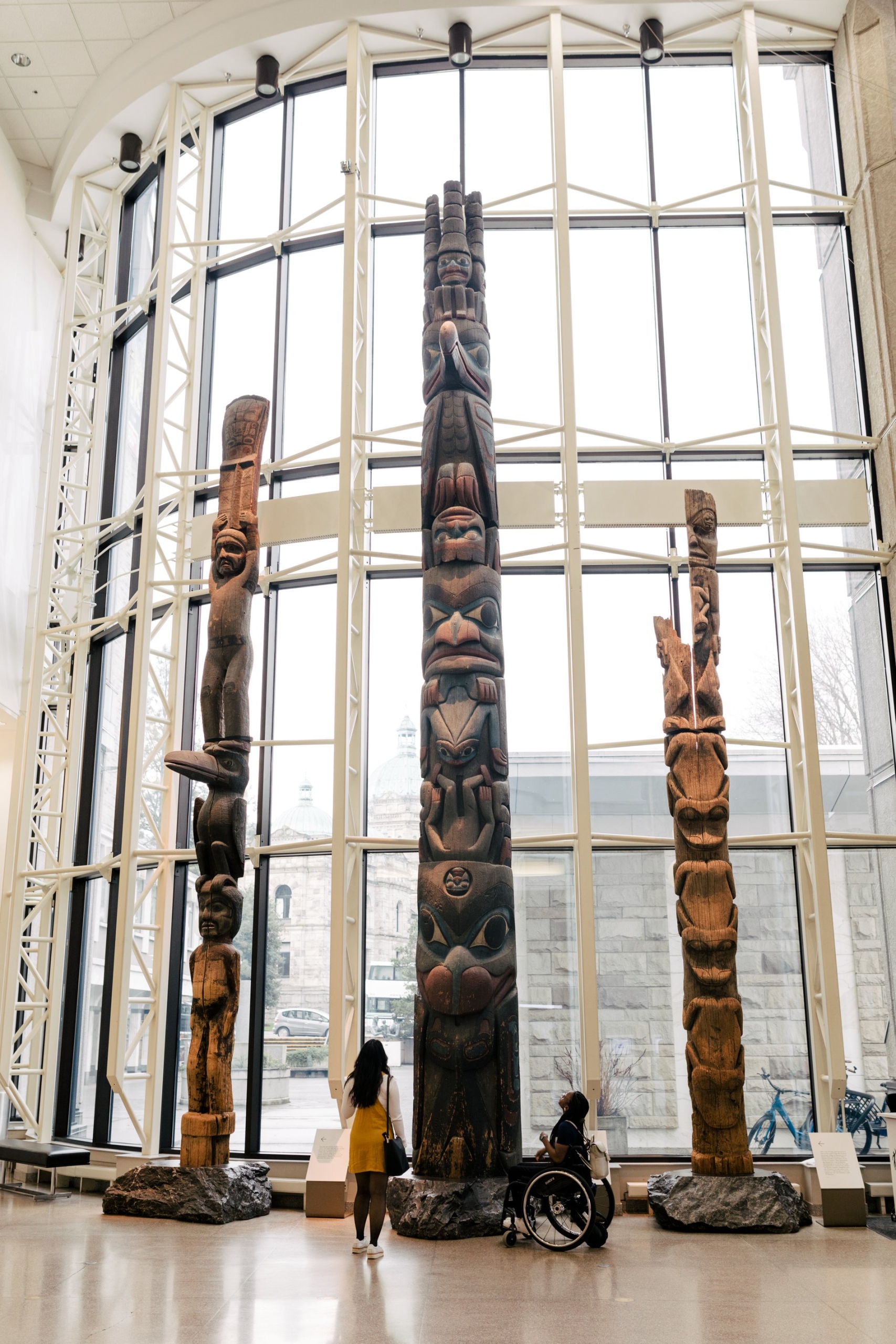 One person in a wheelchair and another stare up at a collection of Totem poles at the Royal BC Museum