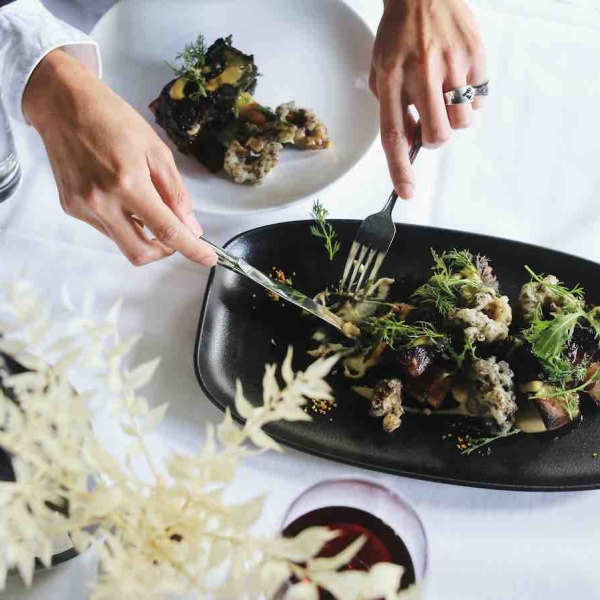 A person uses a knife and fork to cut a piece of cauliflower and salad from a shared plate, only their hands are seen. The table has a white linen table cloth. There is a glass of red wine and a white plant on then table.