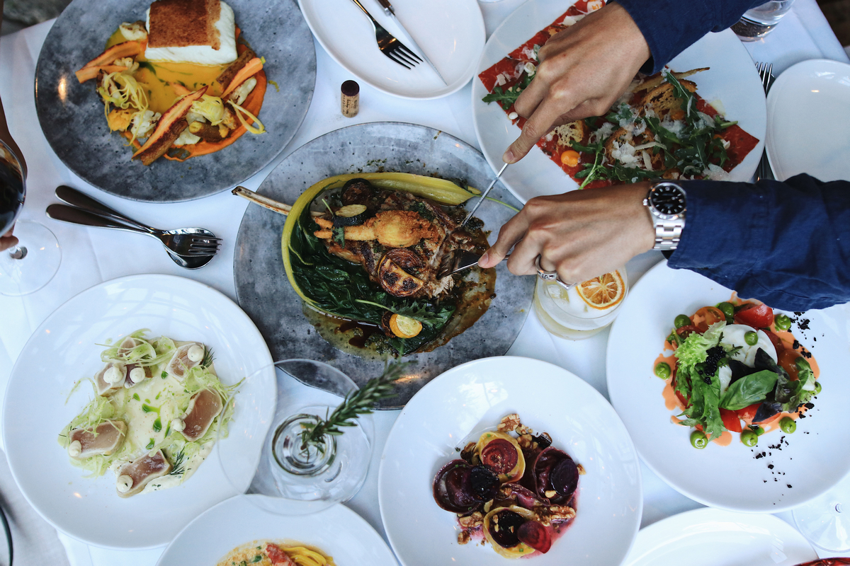 An overhead shot of several dieshes from Caminetto on a table cloth. Hands are seen reaching into the centre dish to cut off a piece of of the dish.