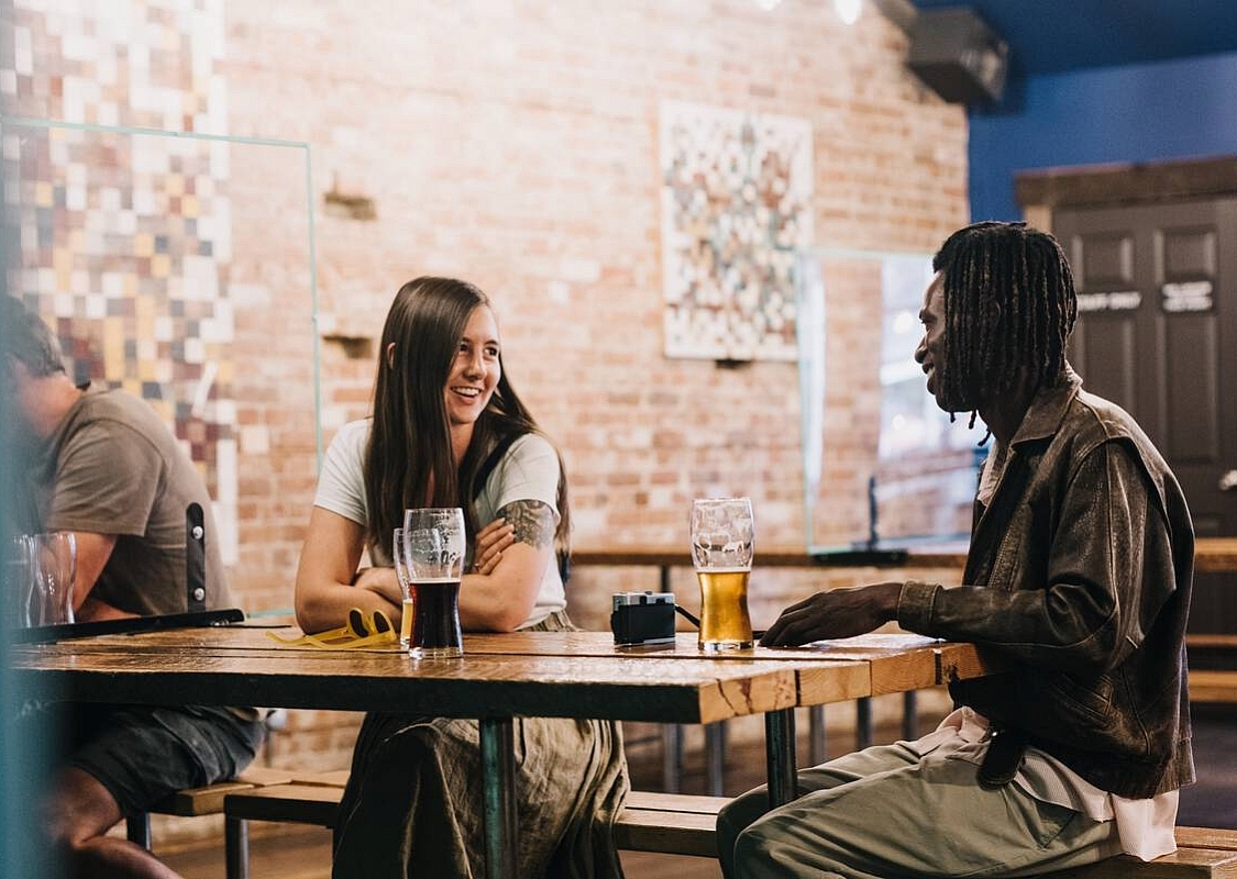 Two people sit at a wooden table enjoying a beer at Backroads Brewing Co. in Nelson. There is a brick wall with artwork behind them and other people off to the side.
