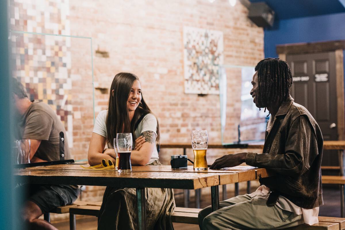 Two people sit at a wooden table enjoying a beer at Backroads Brewing Co. in Nelson. There is a brick wall with artwork behind them and other people off to the side.