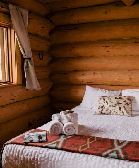 Una cama de matrimonio en el interior de una cabaña de madera en el Big Bar Guest Ranch. La ventana situada junto a la cama está abierta, dejando entrar el aire y la luz.