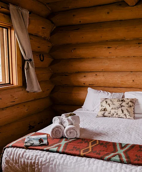 Ein Queensize-Bett in einer Blockhütte auf der Big Bar Guest Ranch. Das Fenster neben dem Bett ist geöffnet und lässt frische Luft und Licht herein.