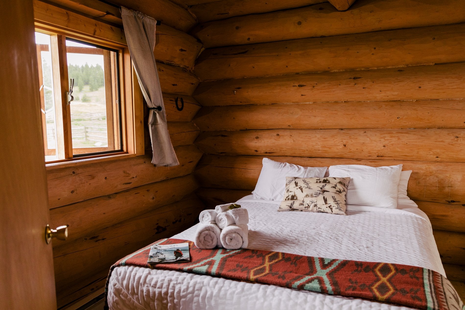 Ein Queensize-Bett in einer Blockhütte auf der Big Bar Guest Ranch. Das Fenster neben dem Bett ist geöffnet und lässt frische Luft und Licht herein.