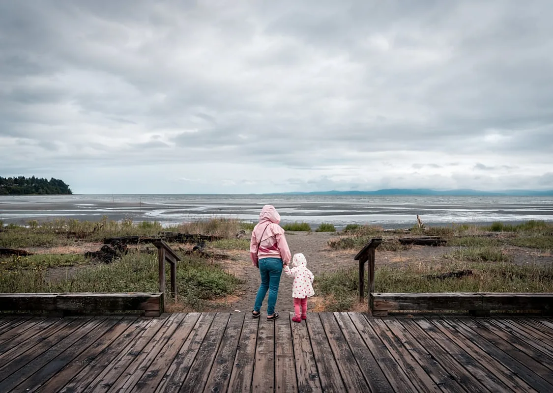 A parent and child stand on a deck looking out at the beach