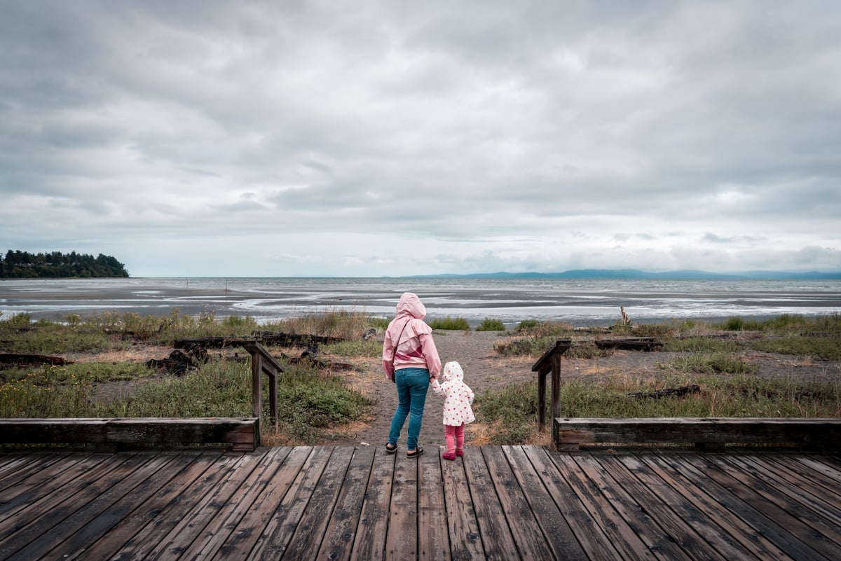 A parent and child stand on a deck looking out at the beach