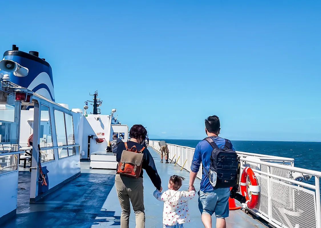 A family walks along the top deck of a BC Ferries boat on a sunny blue ski day. Two parents hold the hand of small child between them.