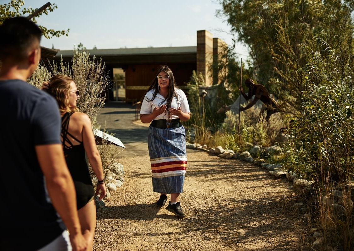 A woman faces the camera, talking to a tour group and leading a cultural tour. A couple watches and listens.