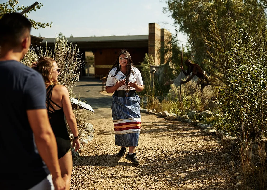 Una mujer mira a la cámara, hablando con un grupo de turistas y guiando un recorrido cultural. Una pareja observa y escucha.