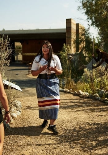 A woman faces the camera, talking to a tour group and leading a cultural tour. A couple watches and listens.