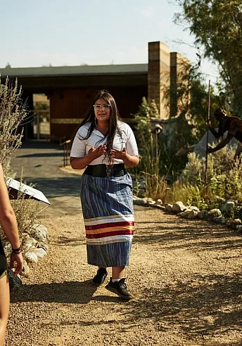 Una mujer mira a la cámara, hablando con un grupo de turistas y guiando un recorrido cultural. Una pareja observa y escucha.
