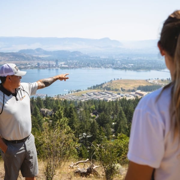 A man points out in the distance towards the lake on a walking tour of the Moccasin Trails. A woman with a pony tail is in the foreground listening.