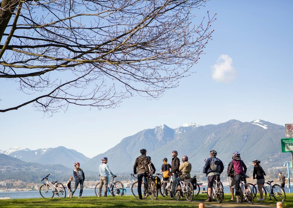 A group of a dozen or so cyclists look across a body of water toward the North Shore Mountains on a clear day.