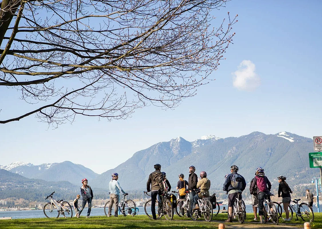 A group of a dozen or so cyclists look across a body of water toward the North Shore Mountains on a clear day.