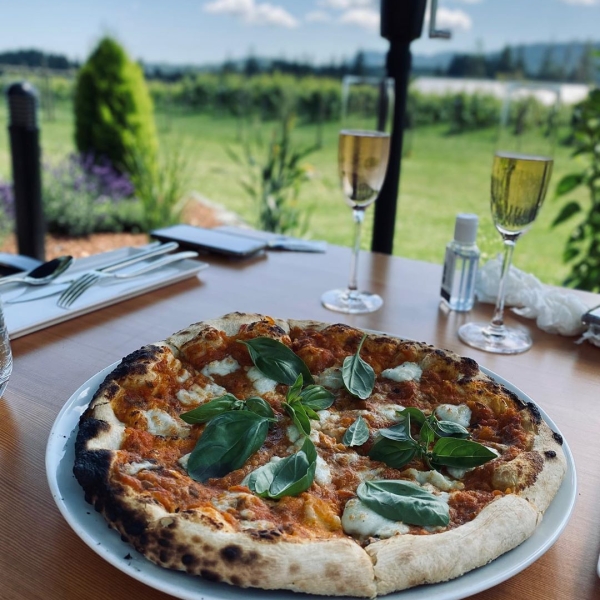 Wood-fired pizza and two glasses of sparkling wine are on a table at Unsworth VIneyards. The table overlooks the vineyard below.