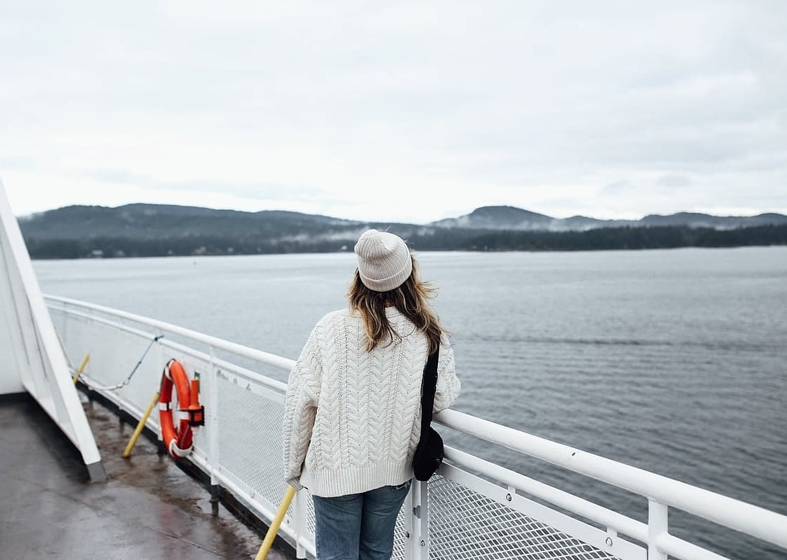 A woman looks out from the ferry deck at the ocean. She is wearing a white sweater and matching toque. The weather is overcast.