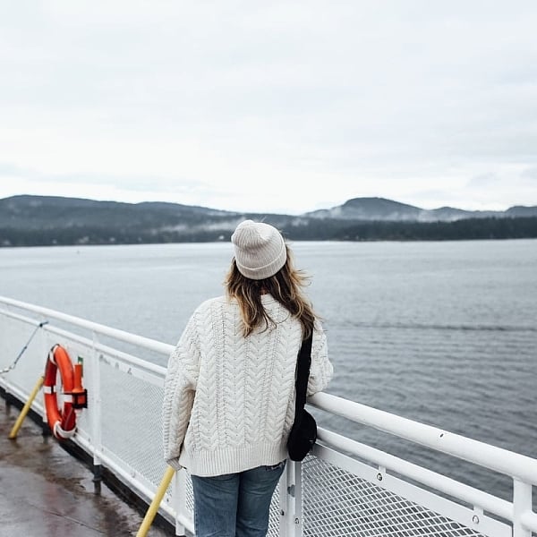 A woman looks out from the ferry deck at the ocean. She is wearing a white sweater and matching toque. The weather is overcast.
