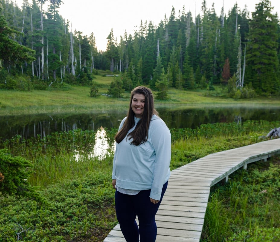 Una persona con una sudadera azul y pantalones negros está parada en un sendero de madera que atraviesa lo que parece ser un área pantanosa con árboles al fondo.