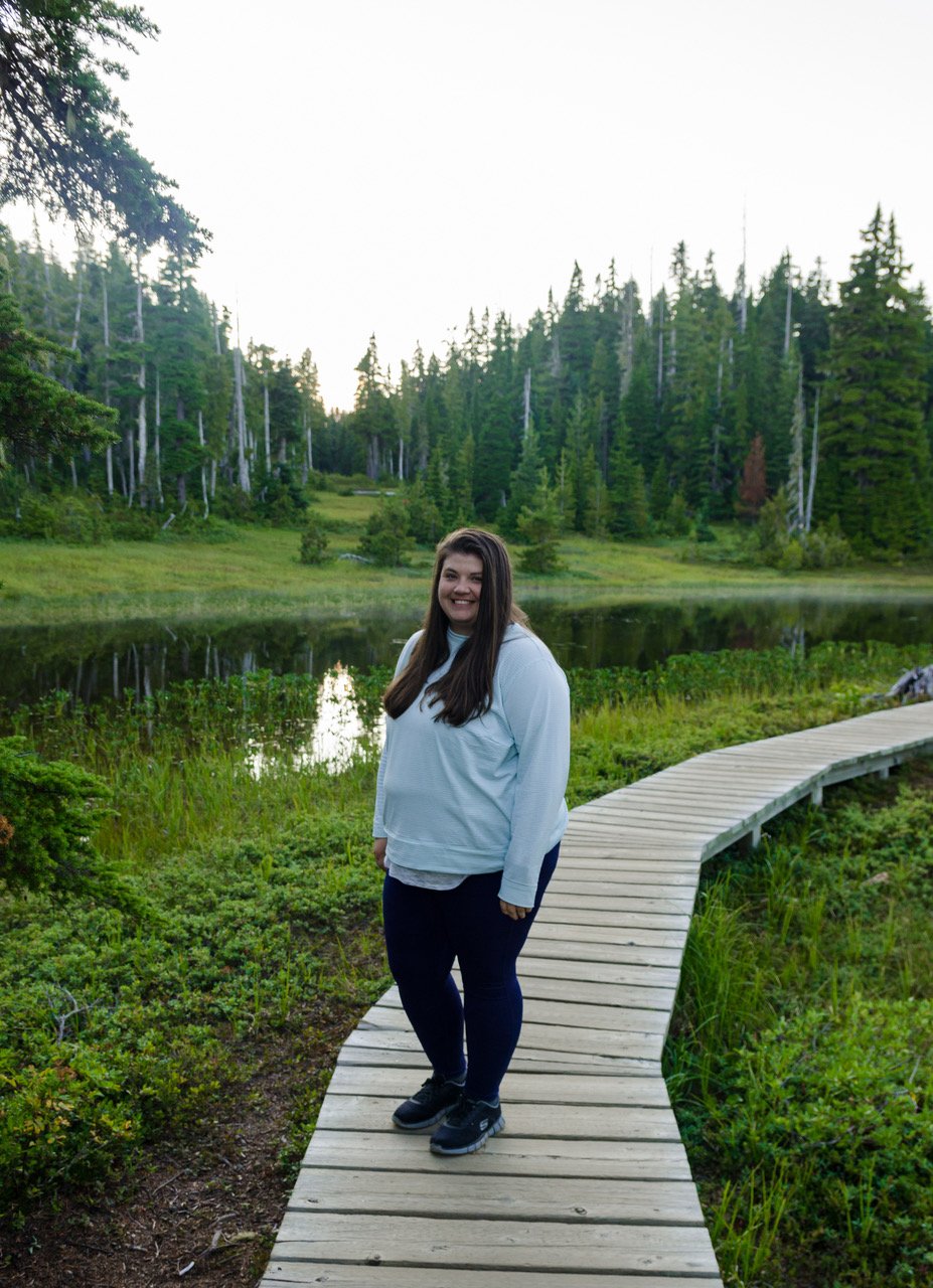 Person in a blue hoodie and black pants stands on a wooden boardwalk trail that runs across what looks to be a marshy area with trees in the background.