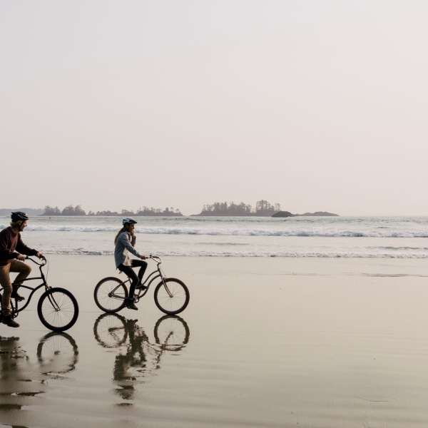 Couple riding bikes along the sand with the ocean and small islands off the coast behind them
