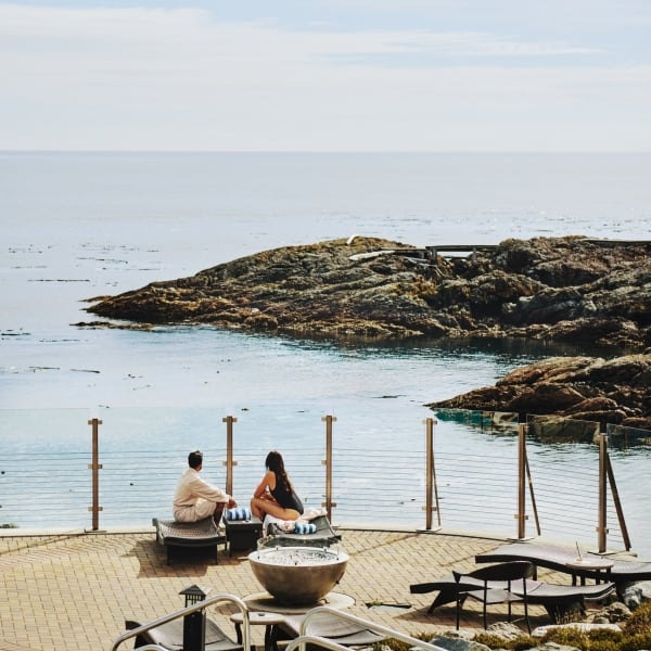 Vista aérea de la terraza de la alberca en el Oak Bay Beach Hotel. Dos personas están sentadas cerca de la barandilla, mirando la costa rocosa.