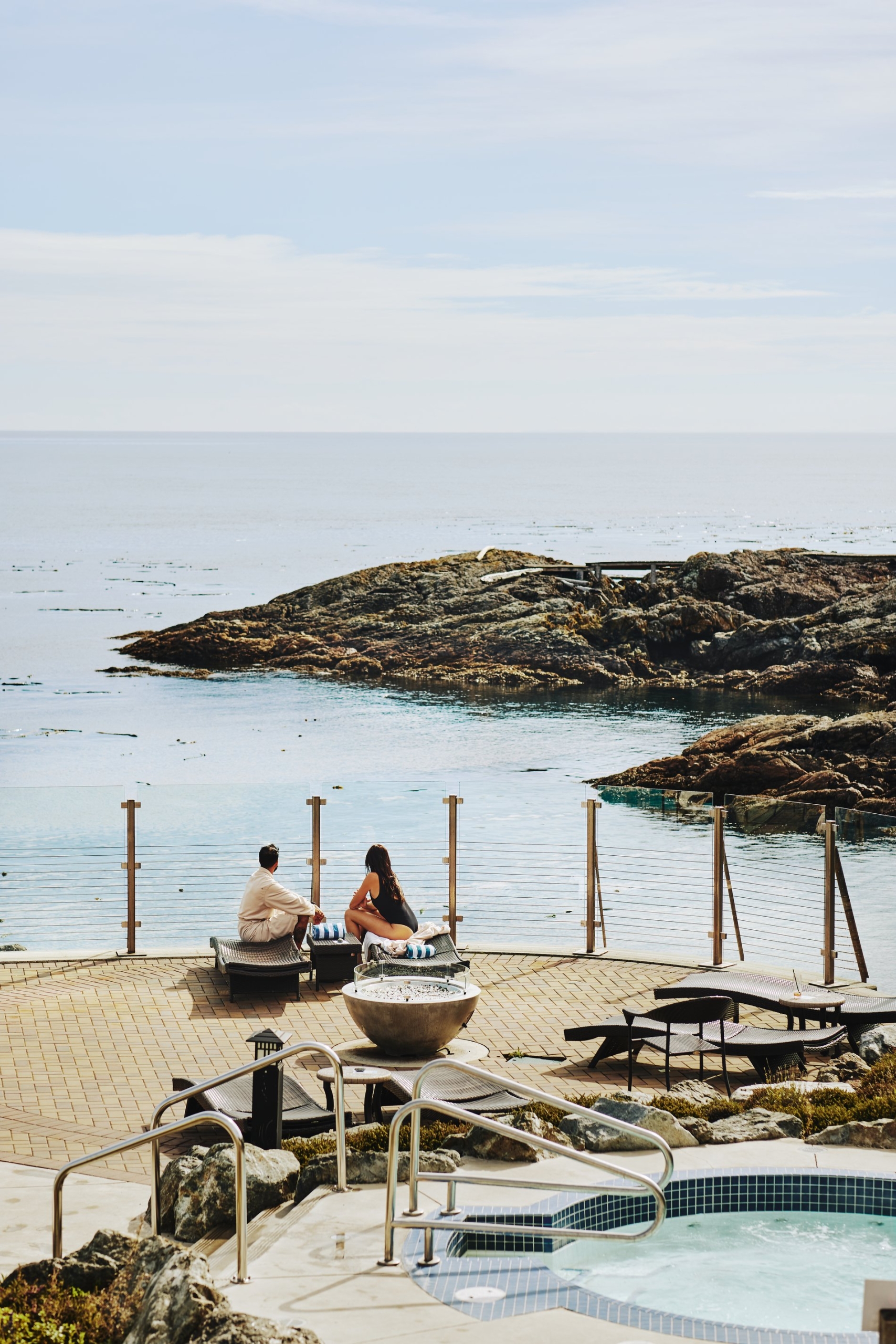 Aerial view of the pool deck at the Oak Bay Beach Hotel. Two people sit near the railing looking out at the rocky shoreline