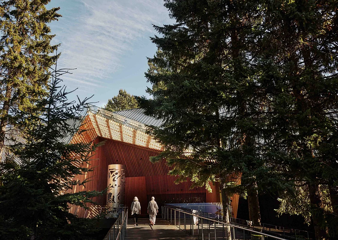 Das Audain Art Museum ist eingebettet in die Bäume in Whistler. Zwei Personen gehen über einen Holzsteg zum hölzernen Eingang.