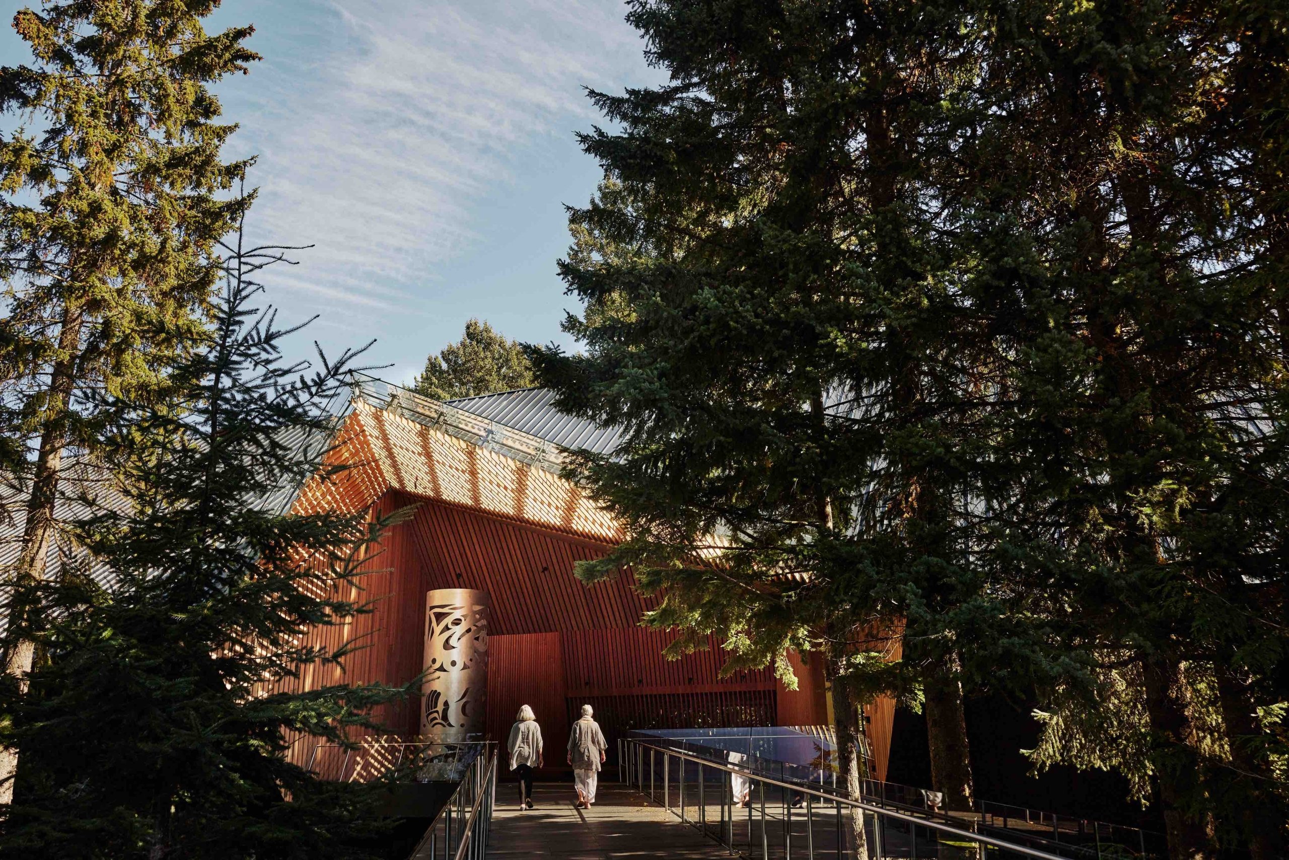The Audain Art Museum is nestled in the trees in Whistler. Two people walk across a boardwalk to the wooden entrance way.