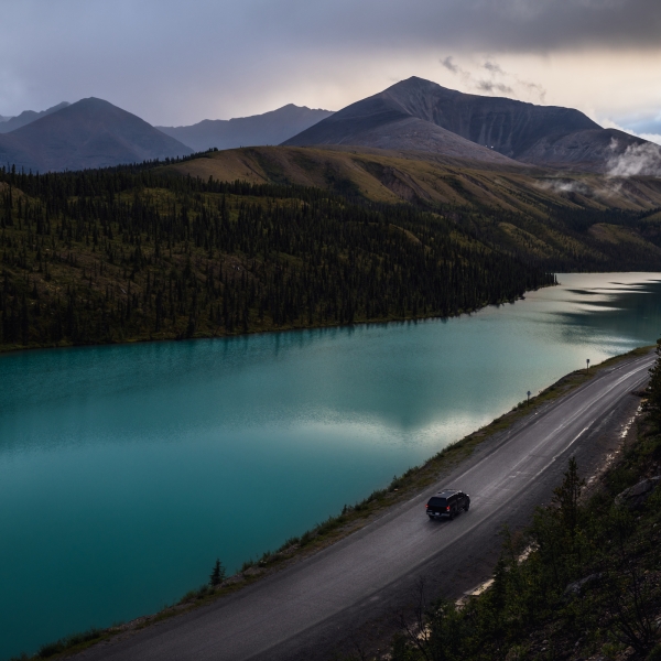 The Alaska Highway winds beside a turquoise lake with mountains in the background.