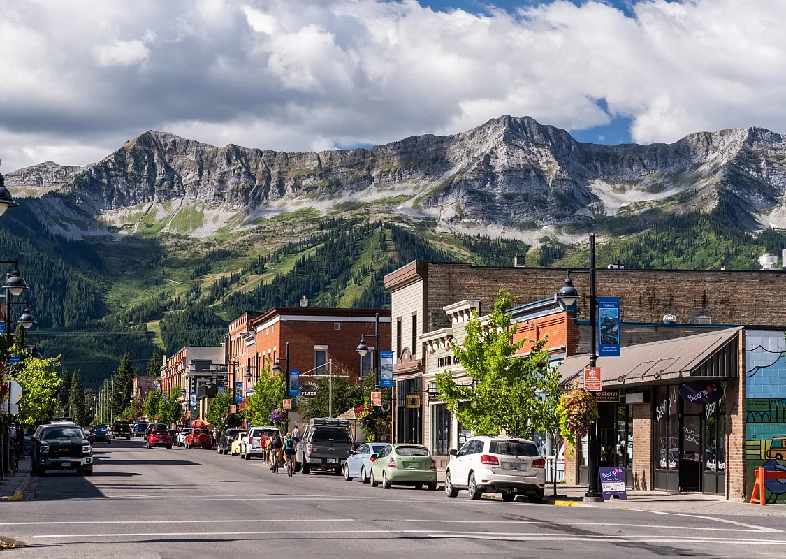 Cars are parked along a quiet street lined with heritage buildings. The sky is cloudy, and a dramatis mountain range is visible in the backdrop of the town.