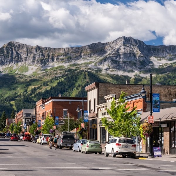Cars are parked along a quiet street lined with heritage buildings. The sky is cloudy, and a dramatis mountain range is visible in the backdrop of the town.