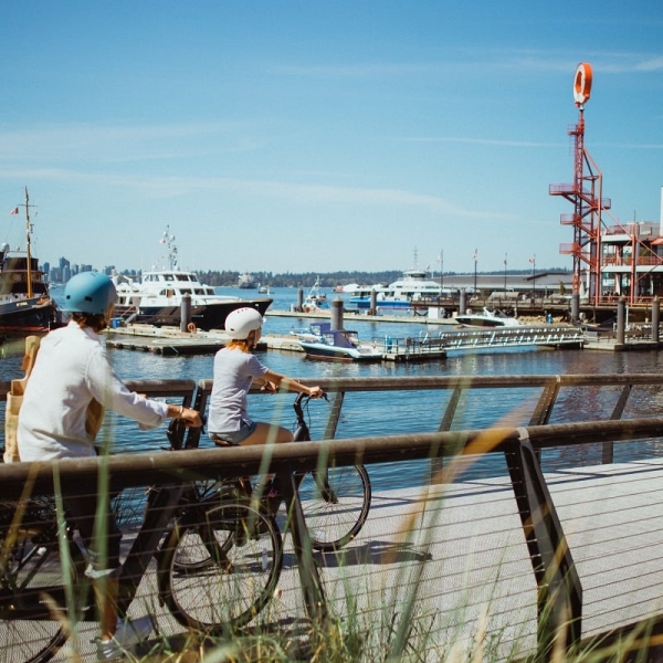 Zwei Radfahrer*innen radeln über eine Brücke am Meer mit den Shipyards im Hintergrund