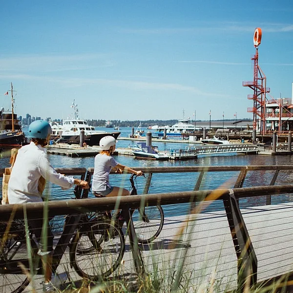 Zwei Radfahrer*innen radeln über eine Brücke am Meer mit den Shipyards im Hintergrund