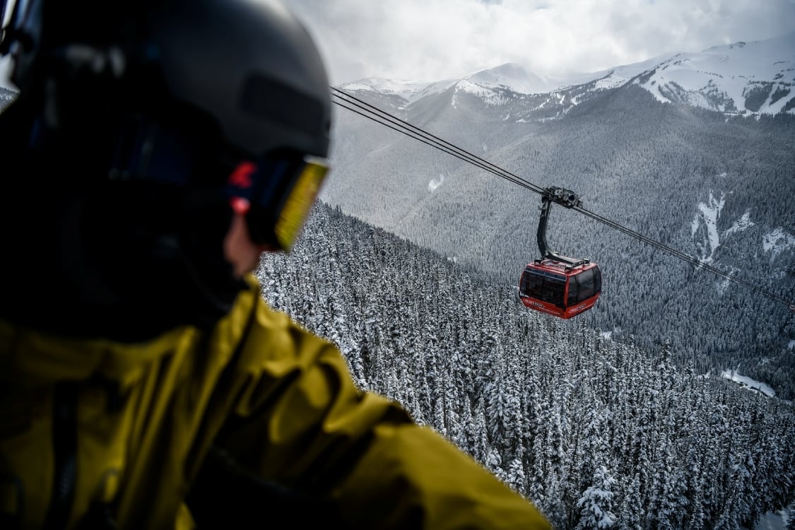 A skier looks down on a passing gondola car that sits high above snowy trees and mountains.