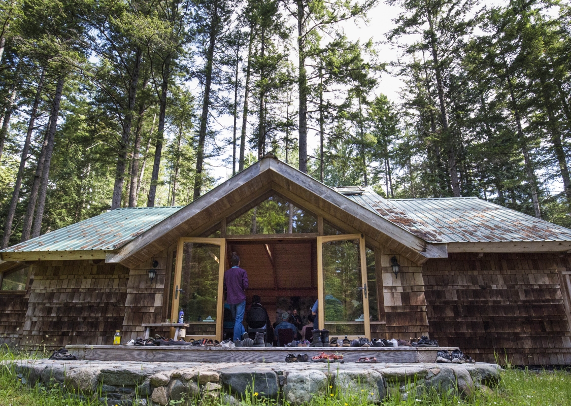 Outside photo of Hollyhock - a wooden lodge with two large glass doors in the centre on Cortes Island. One person stands in the open doorway. Tall trees surround the back of the building, and there is a deck out front.
