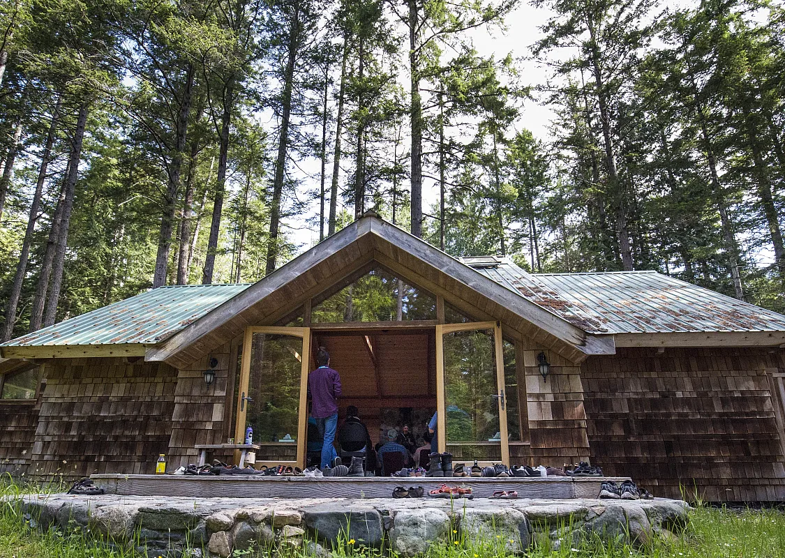 Outside photo of Hollyhock - a wooden lodge with two large glass doors in the centre on Cortes Island. One person stands in the open doorway. Tall trees surround the back of the building, and there is a deck out front.