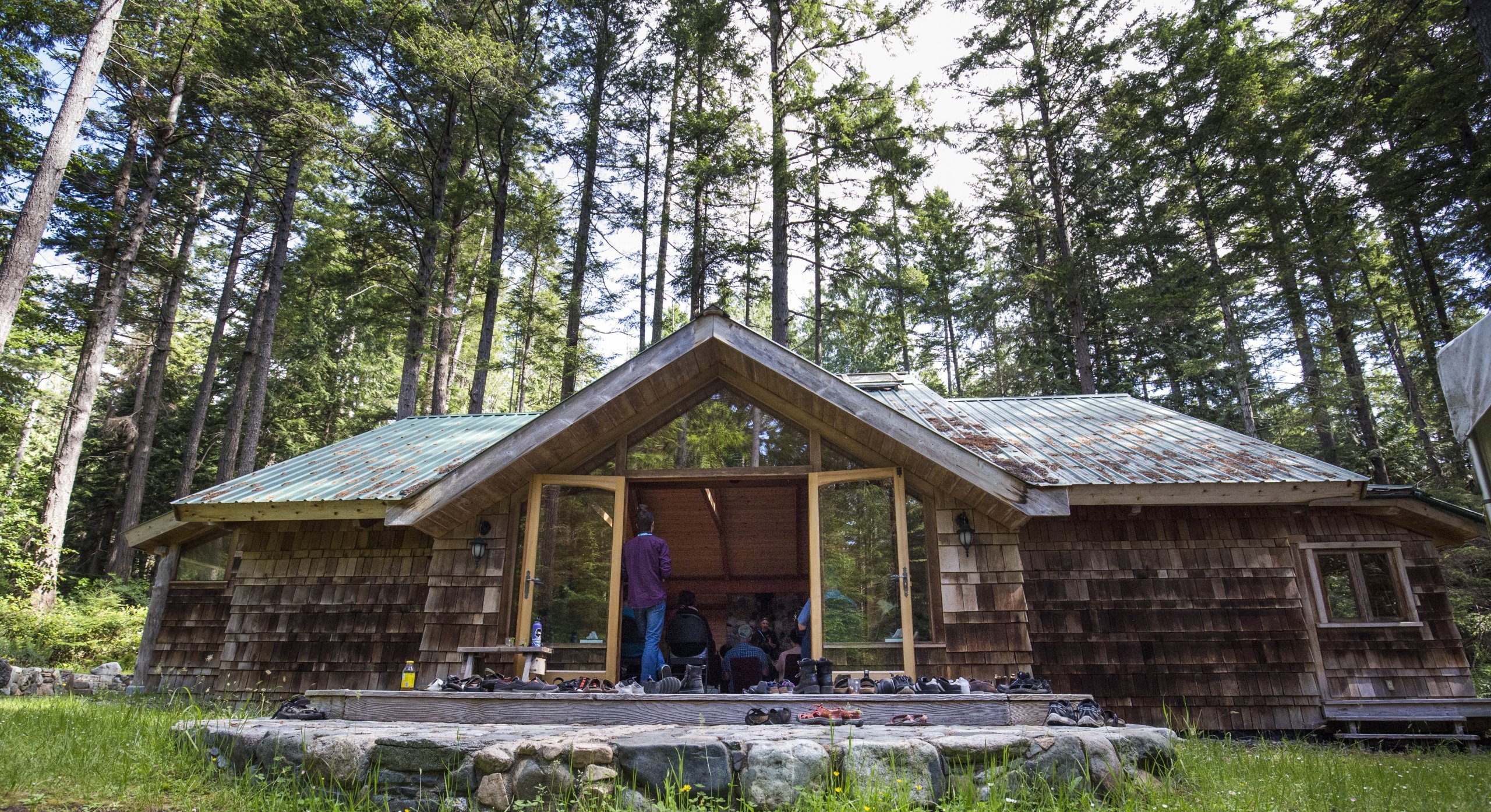 Outside photo of Hollyhock - a wooden lodge with two large glass doors in the centre on Cortes Island. One person stands in the open doorway. Tall trees surround the back of the building, and there is a deck out front.