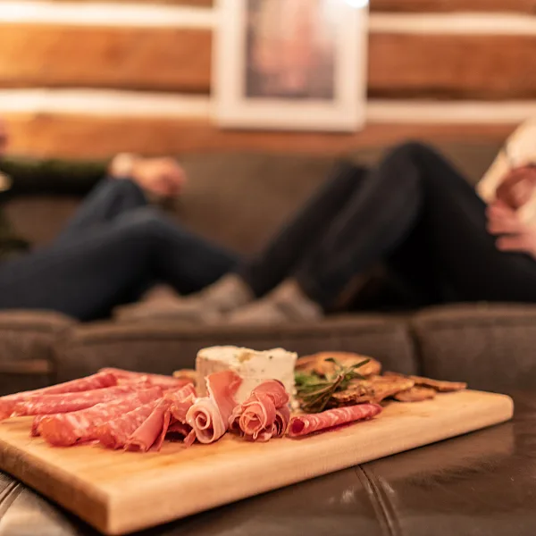 Ein Wurstbrett mit Fleisch und Käse steht auf einem Tisch im Vordergrund, zwei Frauen sitzen auf einer Couch im Hintergrund.m