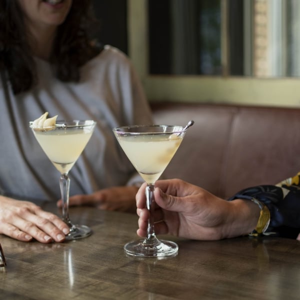 Two people sit at a table holding martini glasses with a yellow beverage. Eyeglasses sit on the table.