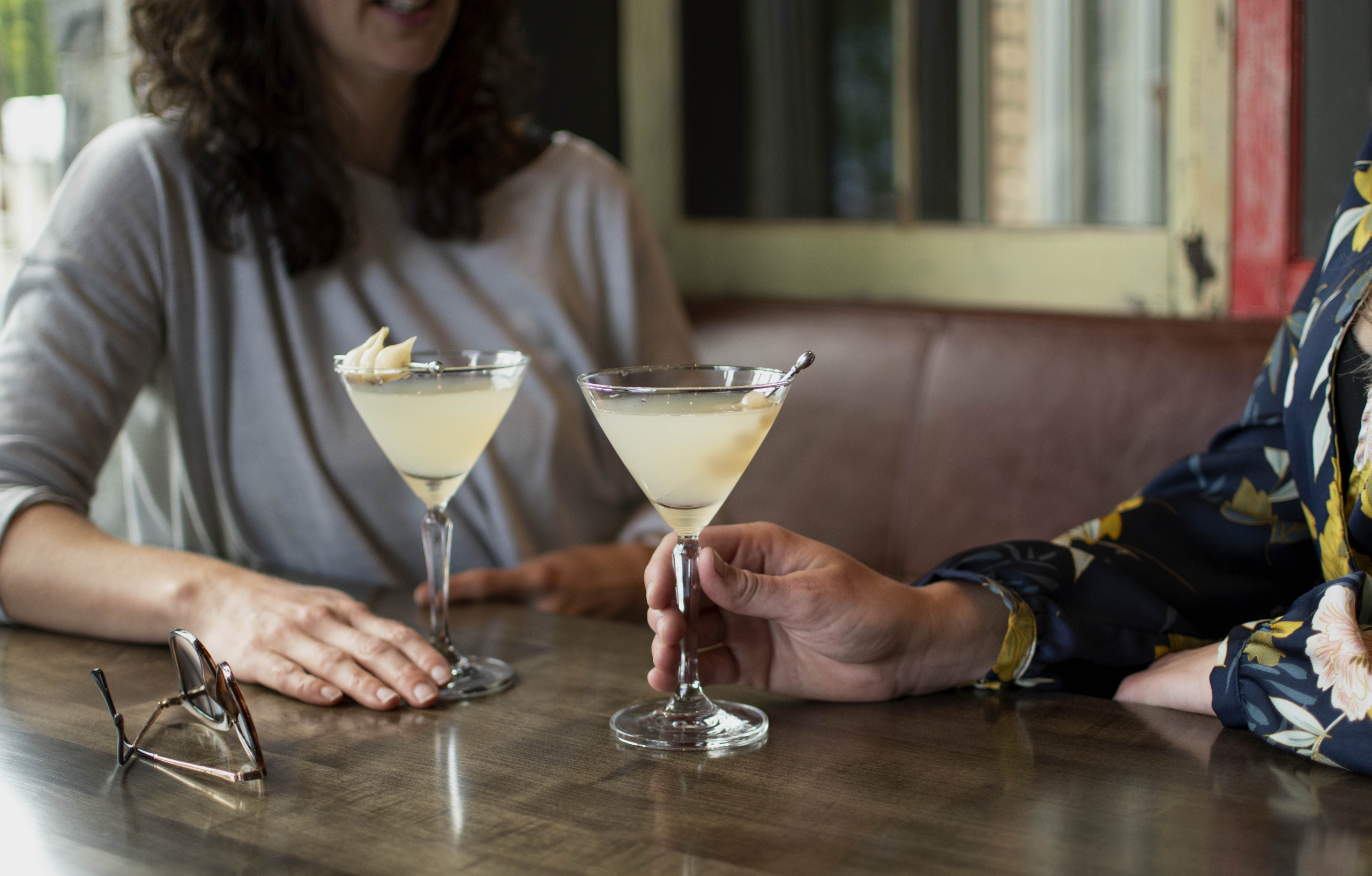 Two people sit at a table holding martini glasses with a yellow beverage. Eyeglasses sit on the table.