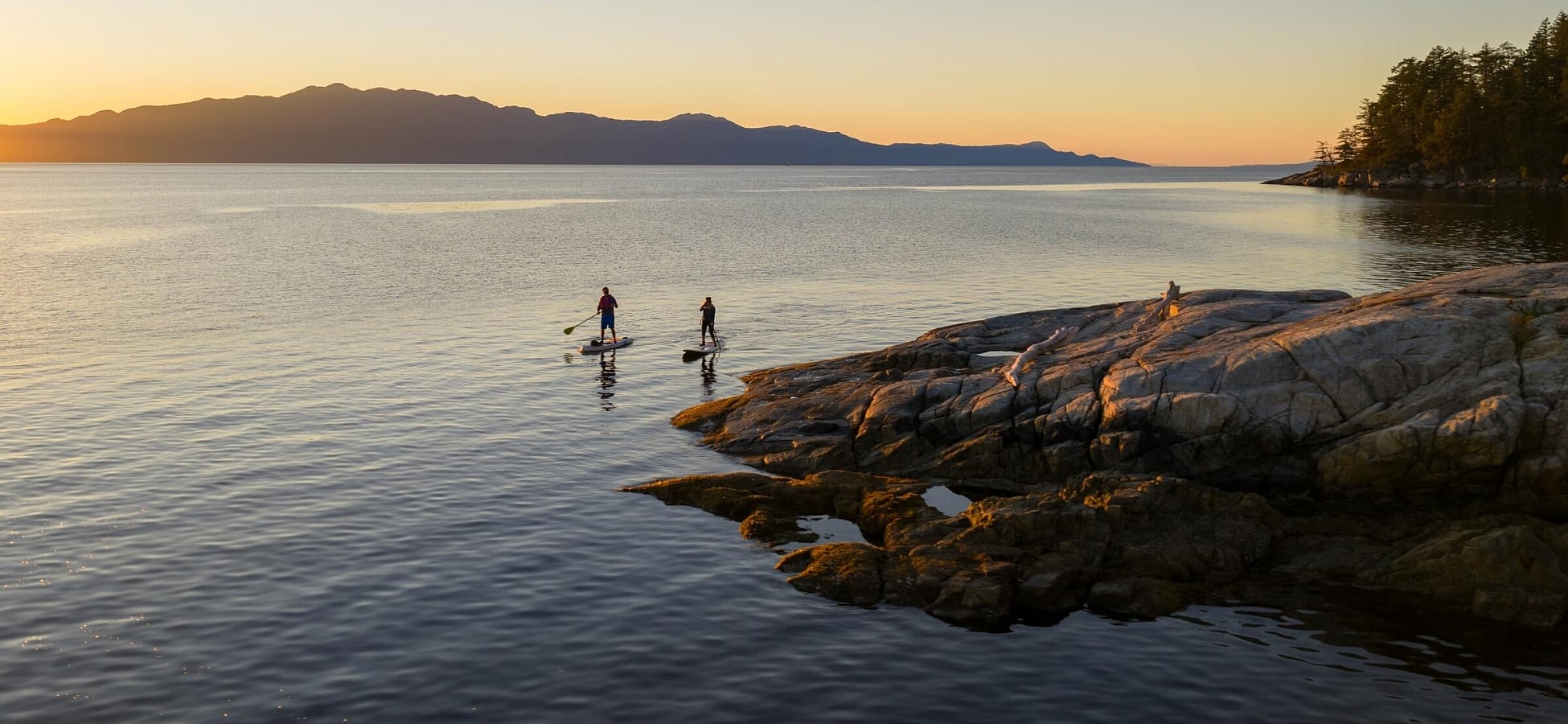 Two people paddleboard off a rocky shoreline.