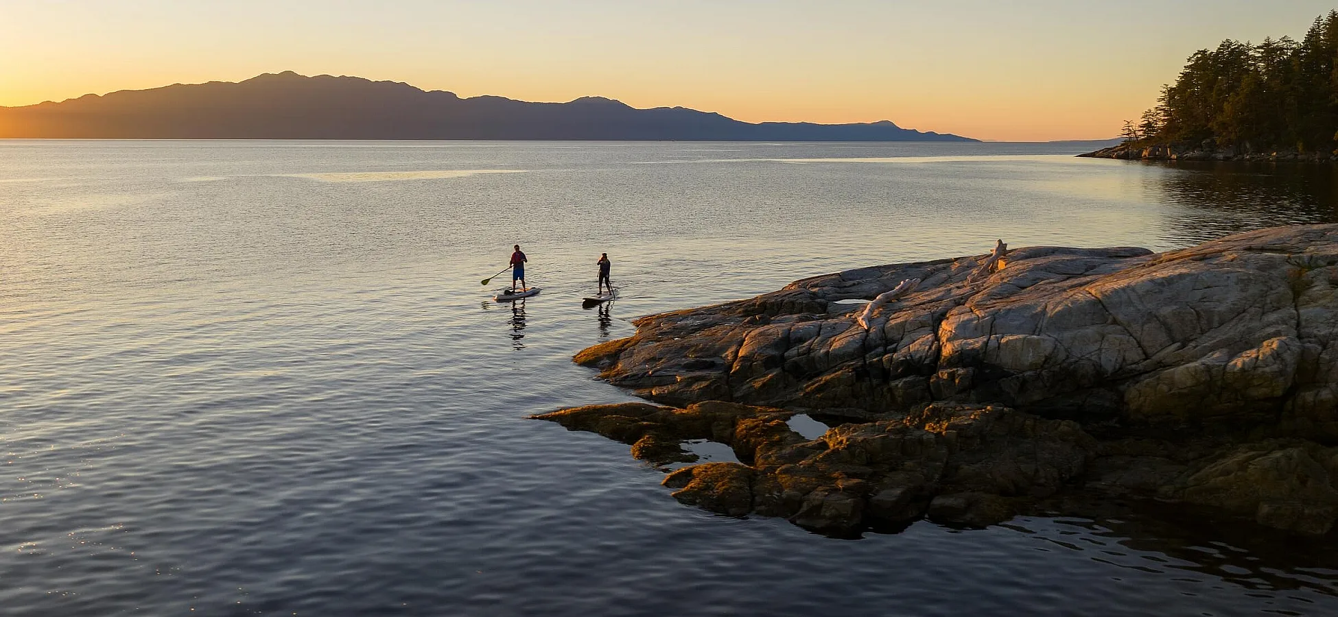 Two people paddleboard off a rocky shoreline.