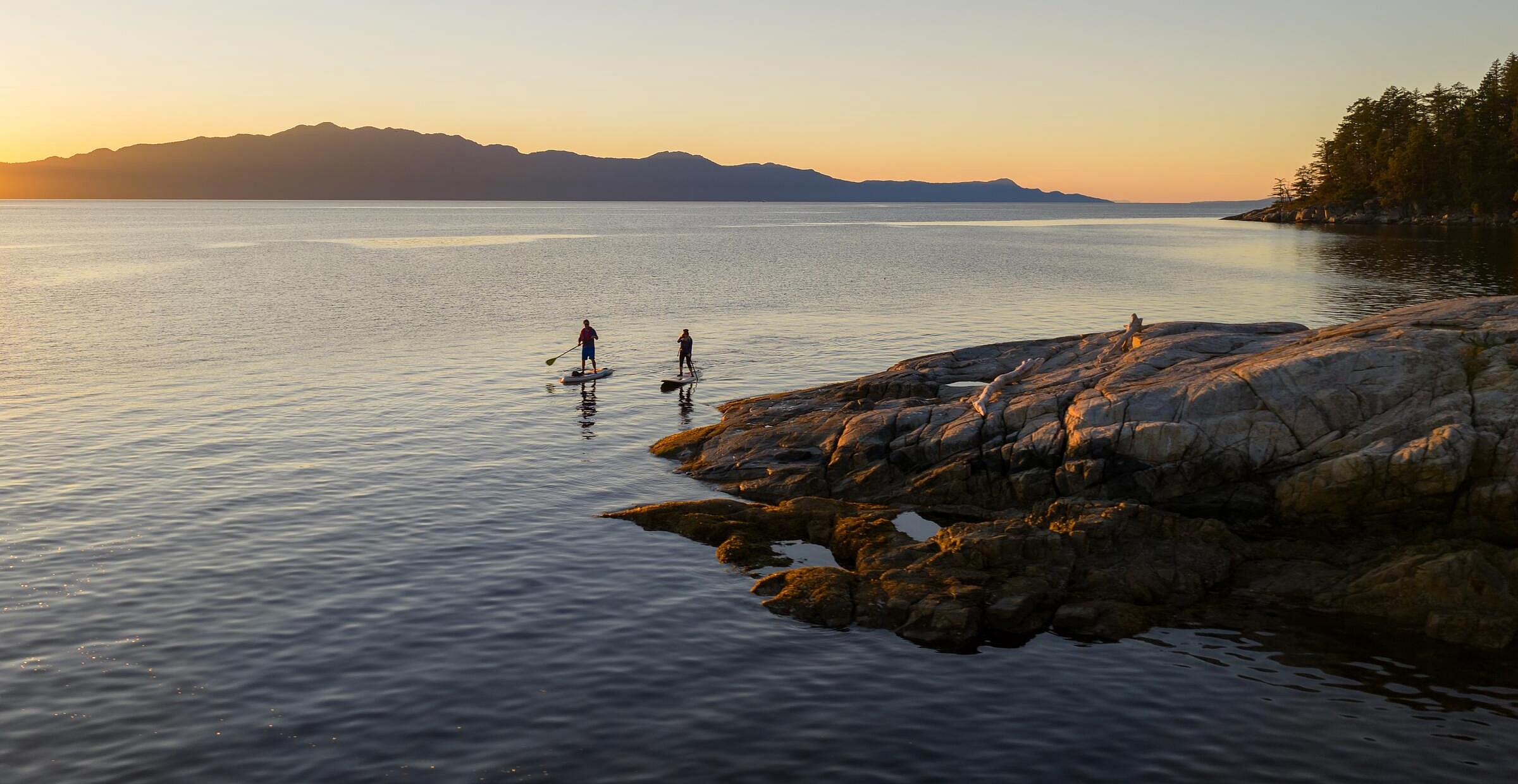 Two people paddleboard off a rocky shoreline.
