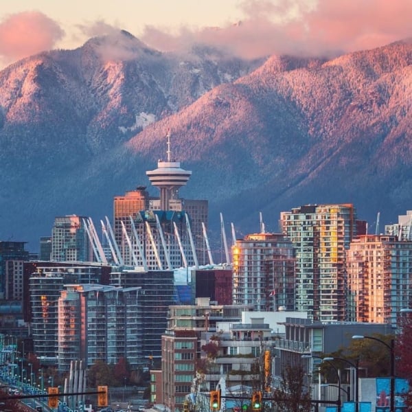 Tall buildings in downtown Vancouver stand in front of snowy mountains in the distance. The sunset is casting a pink hue onto the mountains.