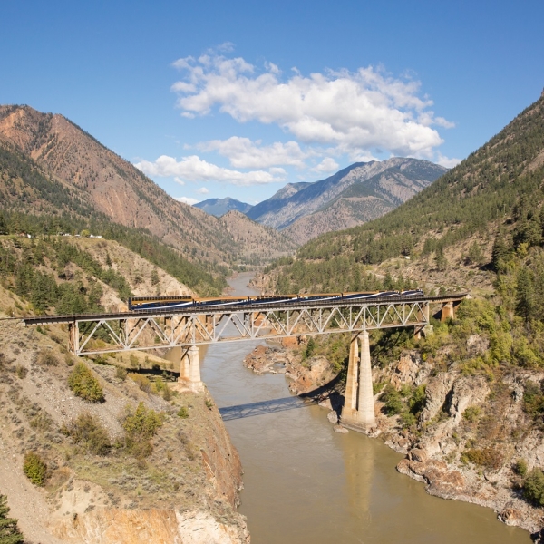 A train crosses a short bridge that crosses the brown, silty Fraser River. On the sides and in the distance, mountains with sparse vegetation.