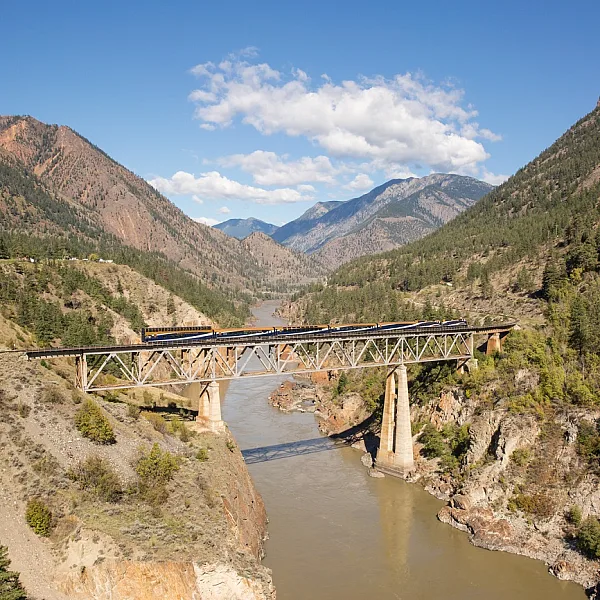 A train crosses a short bridge that crosses the brown, silty Fraser River. On the sides and in the distance, mountains with sparse vegetation.