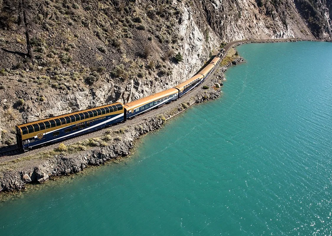 On the left a train travels close to a vibrant turquoise lake with a rocky cliff to the left of the train