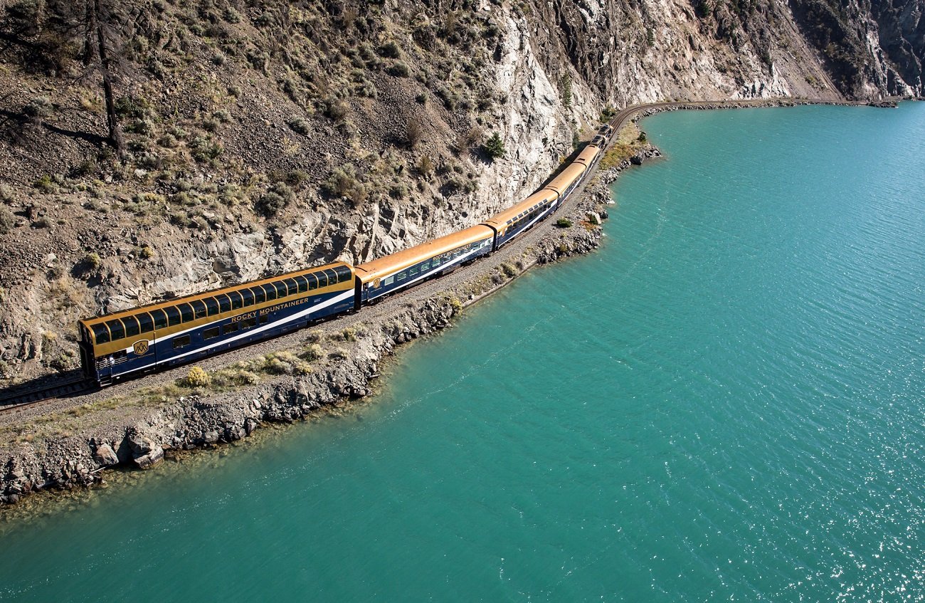 On the left a train travels close to a vibrant turquoise lake with a rocky cliff to the left of the train