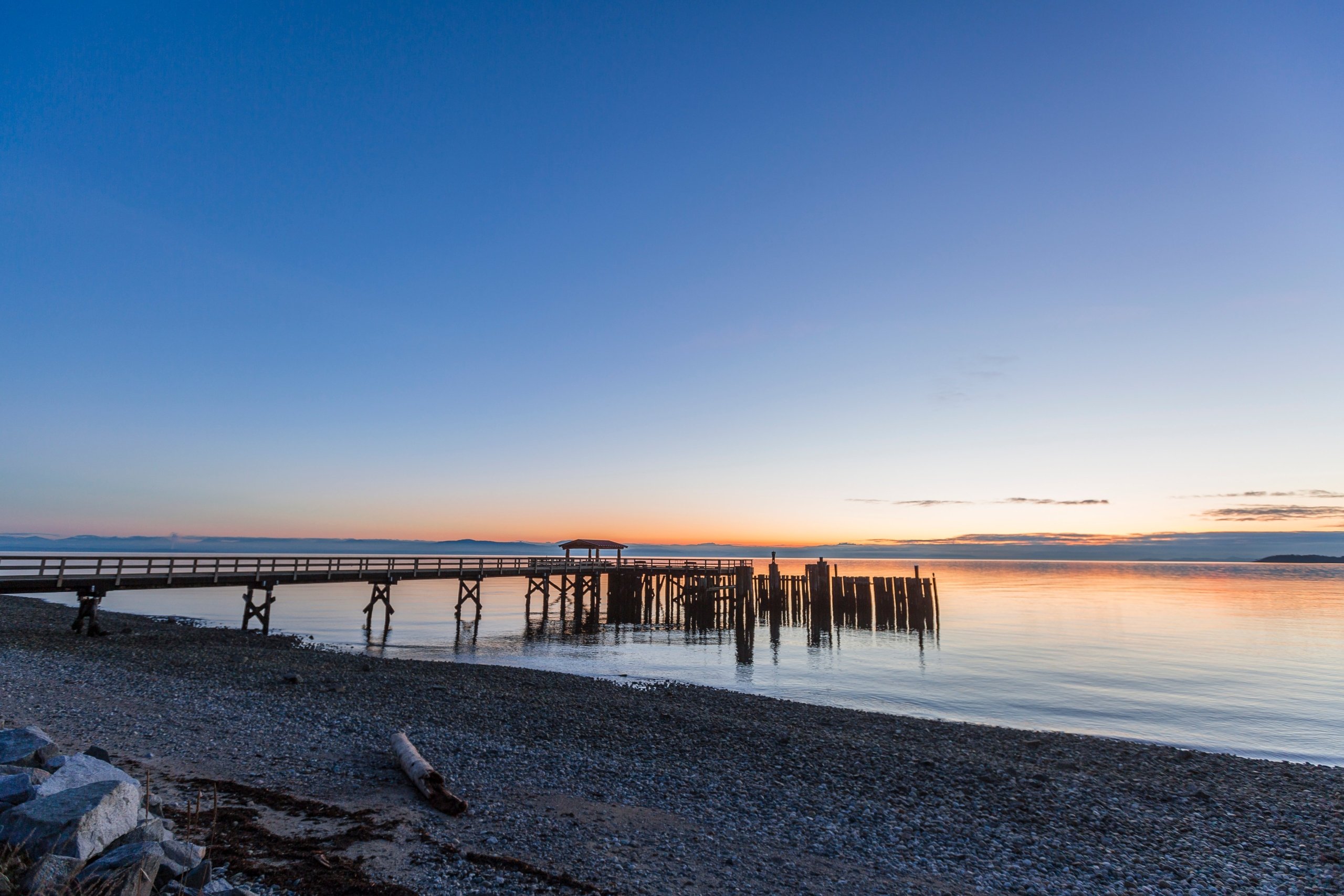 A pier over the water in the sunset with a sandy beach in the foreground.