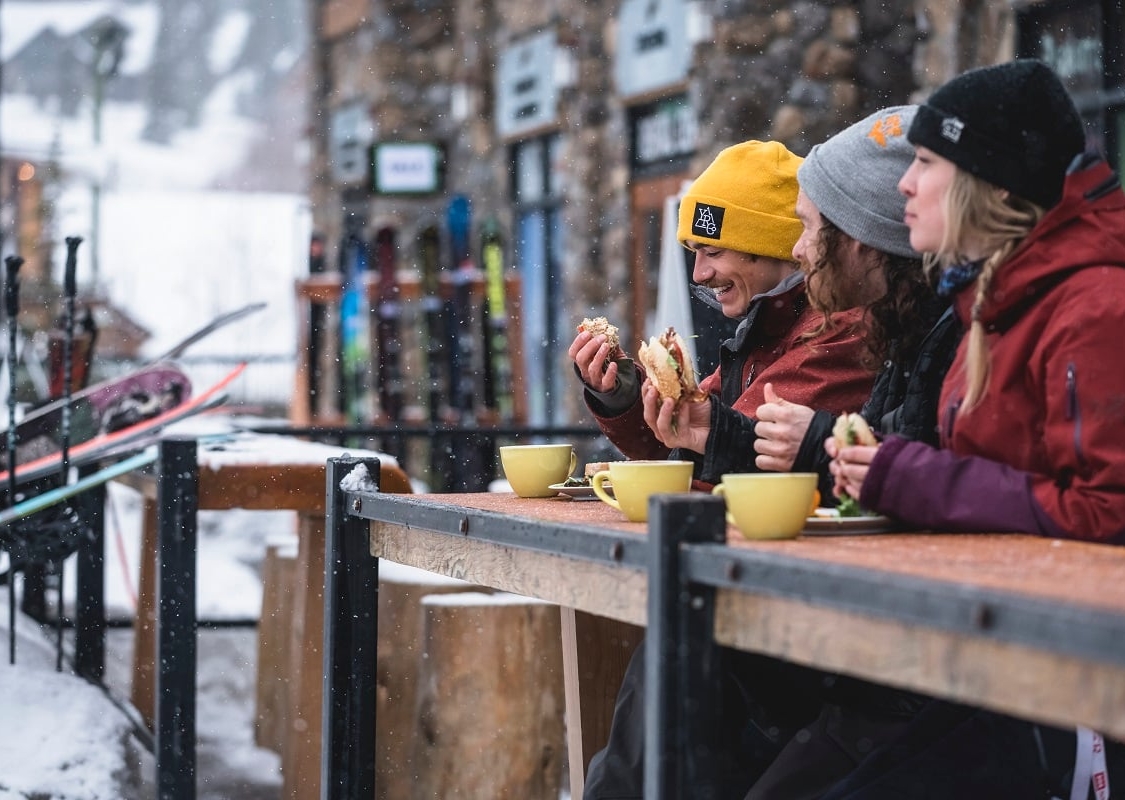 Tres personas sentadas en un patio al aire libre comiendo y bebiendo en invierno. Está nevando suavemente y están vestidos con ropa de invierno.